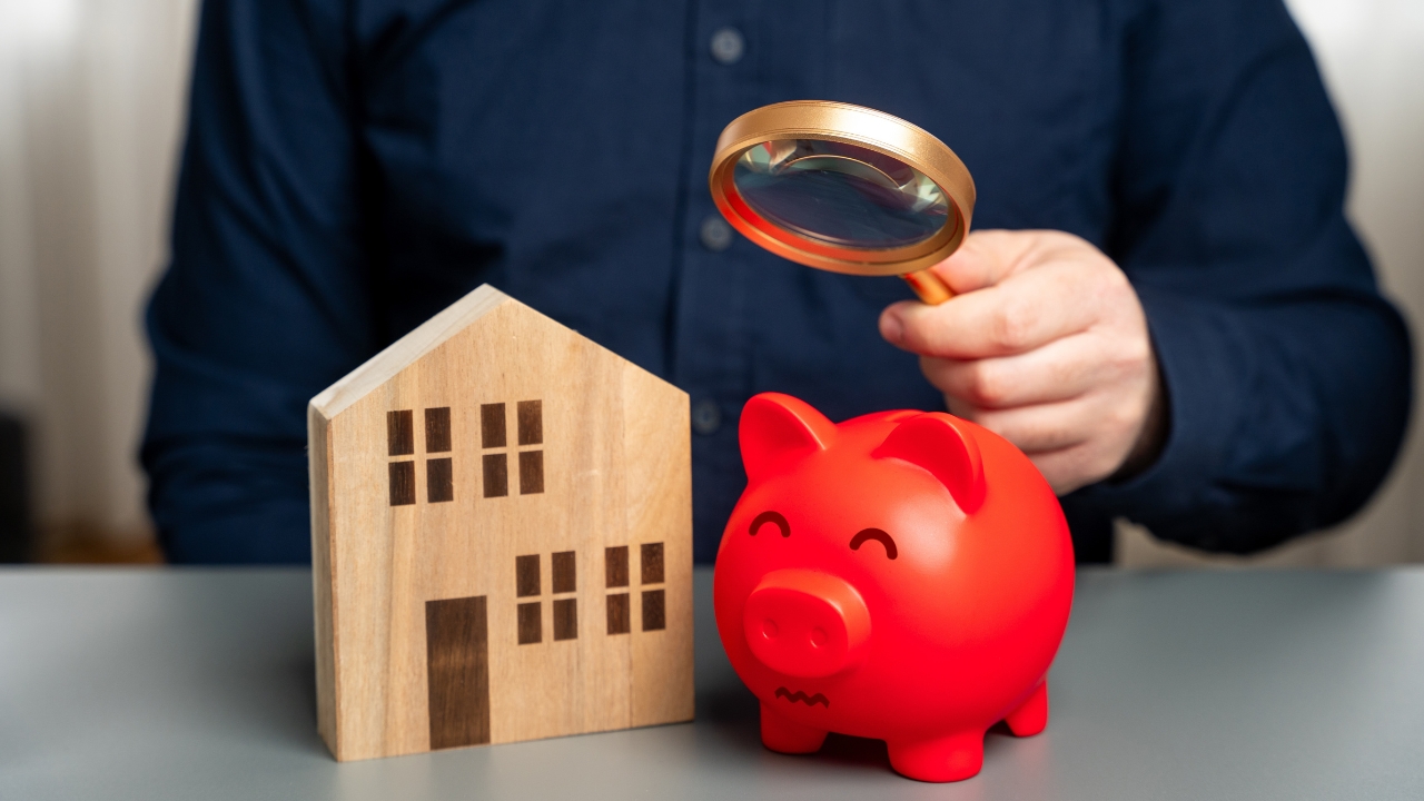 Person holding a magnifying glass near a piggy bank and a small house model