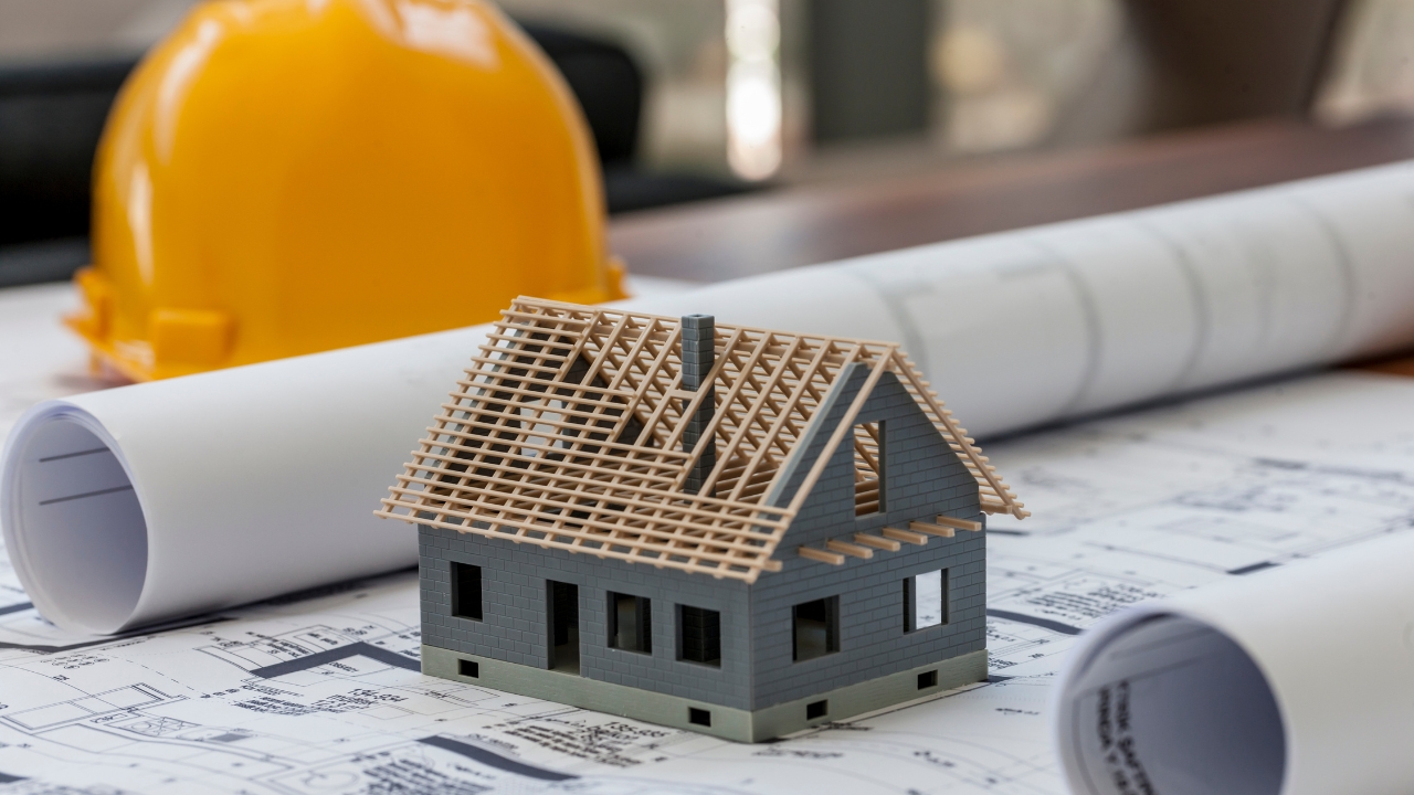 Model house with exposed roof frame placed on construction plans next to rolled blueprints and a hard hat