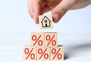 Hand places a house block above mortgage rate symbols for a fixed vs adjustable mortgage comparison