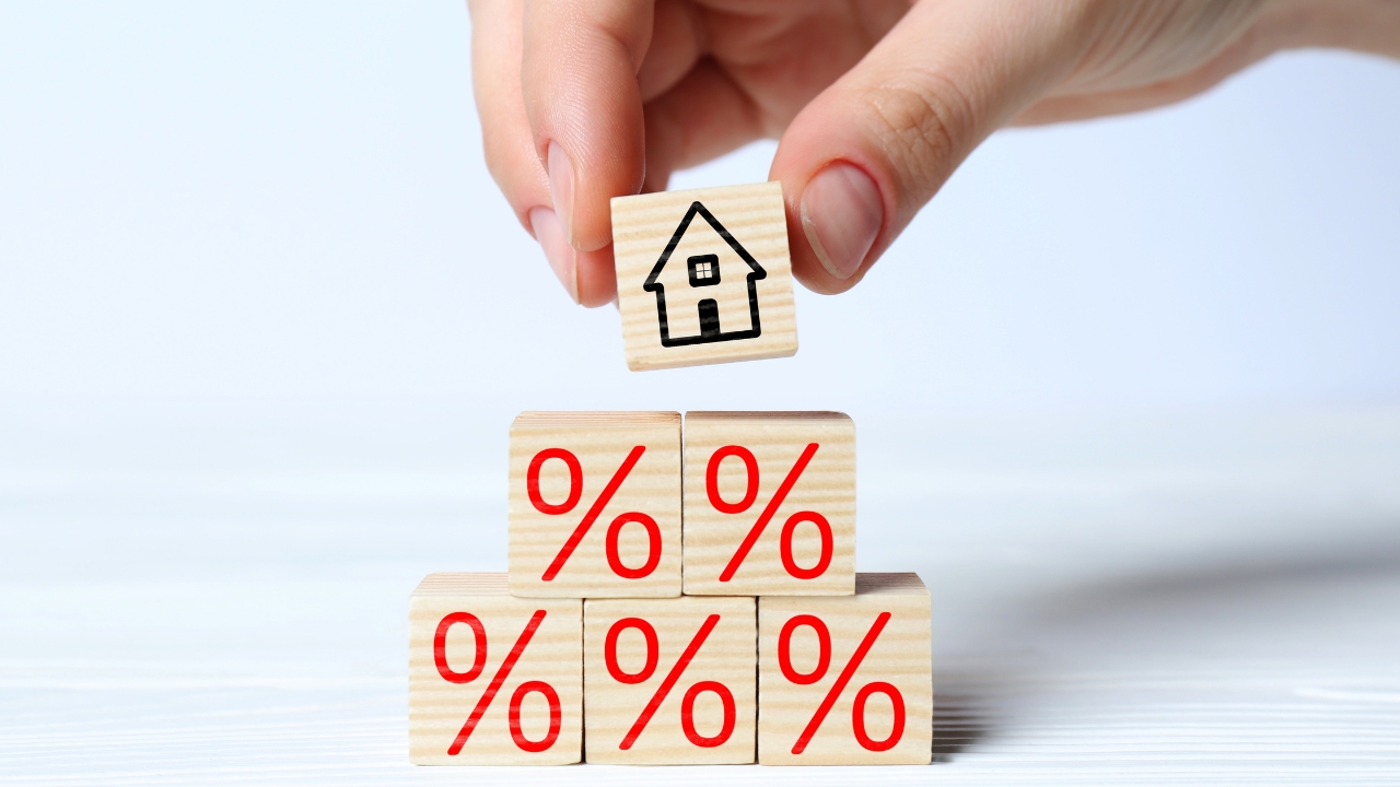 Hand places a house block above mortgage rate symbols for a fixed vs adjustable mortgage comparison