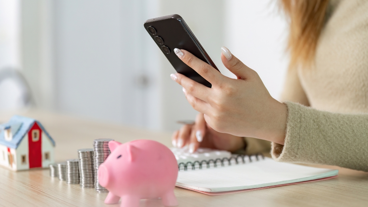 A woman uses her phone to review a home budget beside coins, a notebook, and a piggy bank