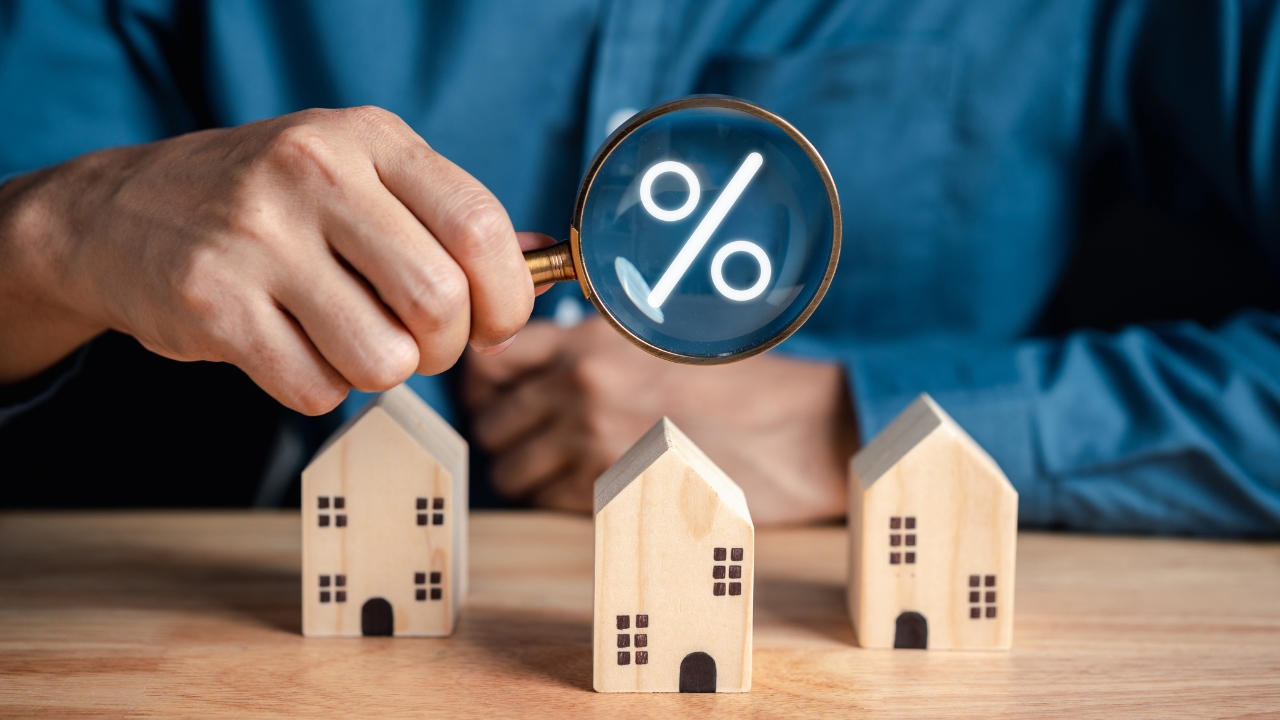 A person checks mortgage rates with a magnifying glass above small wooden houses