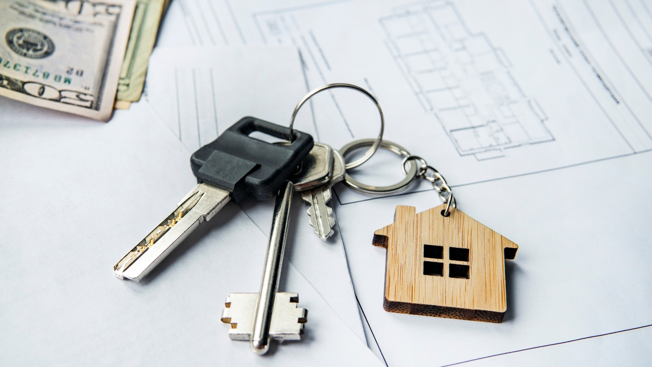House keys and a small wooden home sit on property plans beside cash